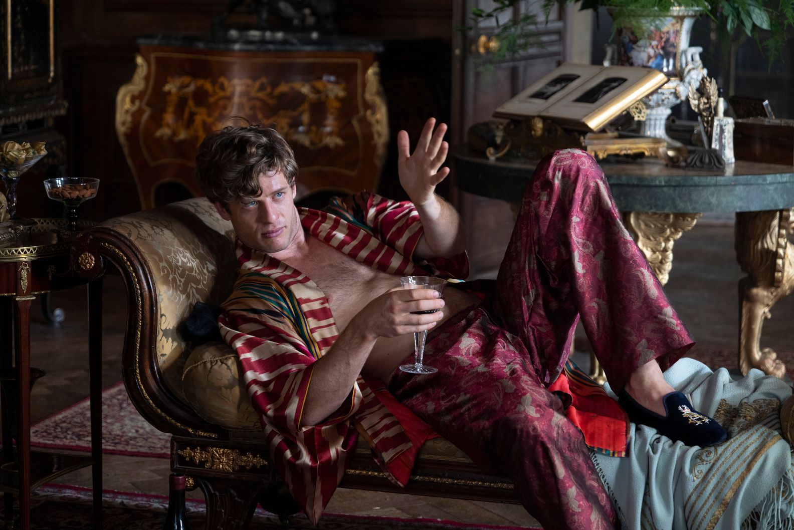 A character lays back on a lounge while bare chested and wearing a silk robe. 