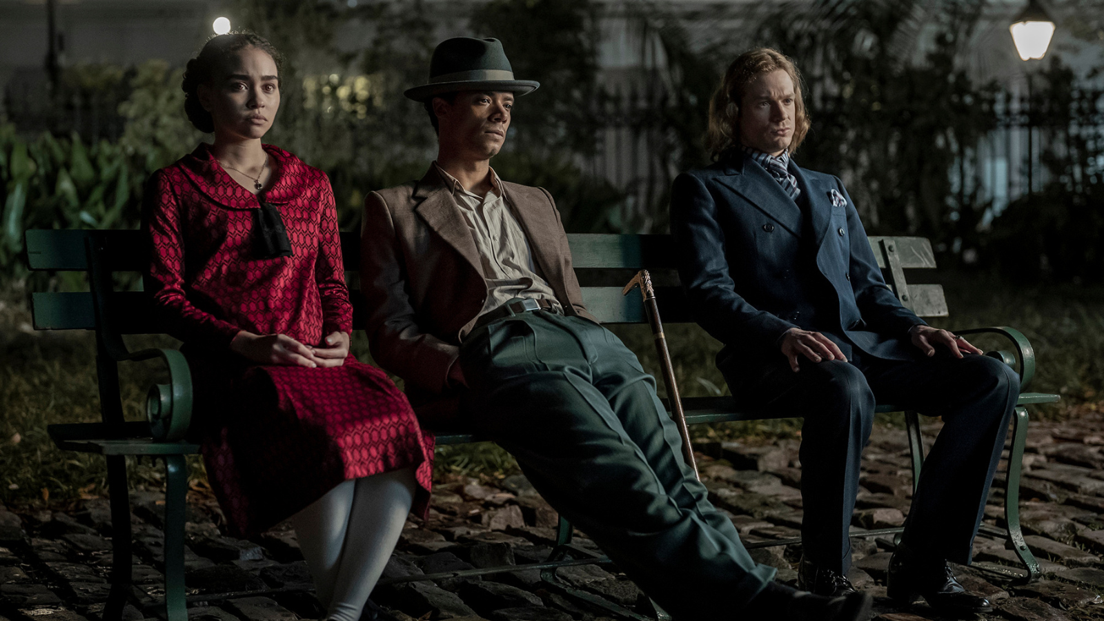 Claudia in a red dress, Louis in browns and greens, and Lestat in a blue suit as they sit on a bench in Jackson Square